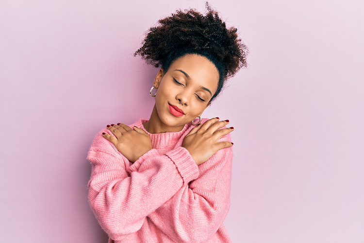 Woman hugging herself in a pink sweater and a purple background. Befriending Your Body