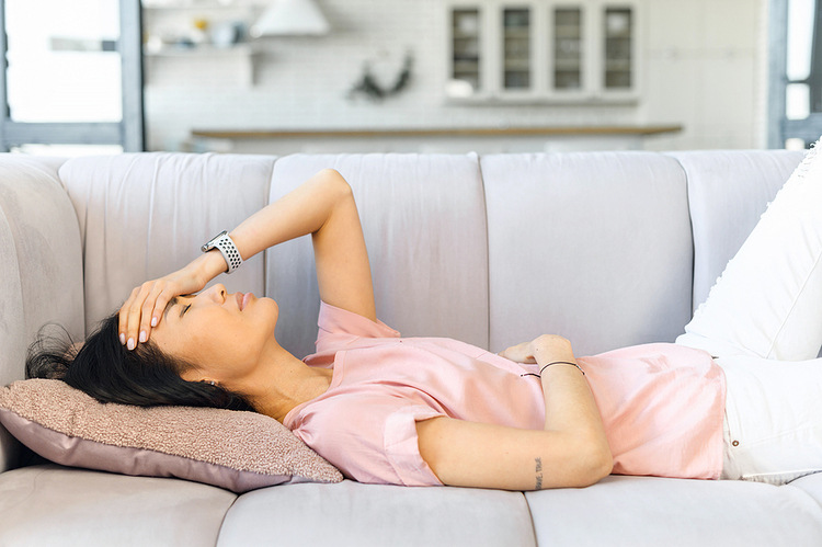 Binge eat. Woman lying on a grey couch wearing a pink shirt. She's holding her stomach and in pain.