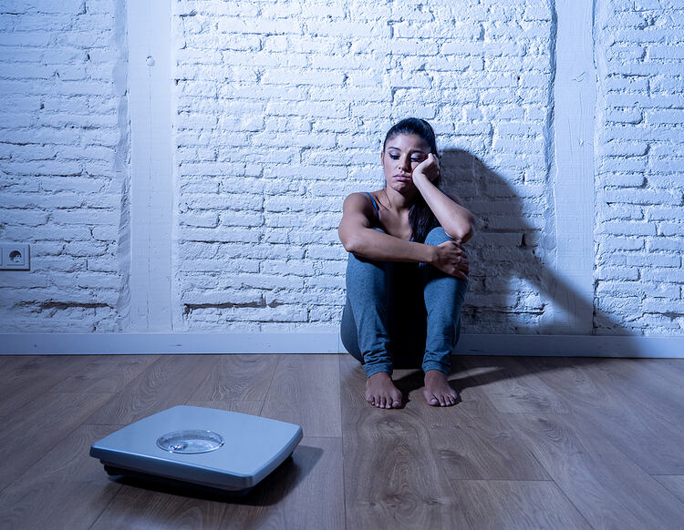 Sad Woman Looking At Scale while sitting on the floor. Eating Disorder Recovery.
