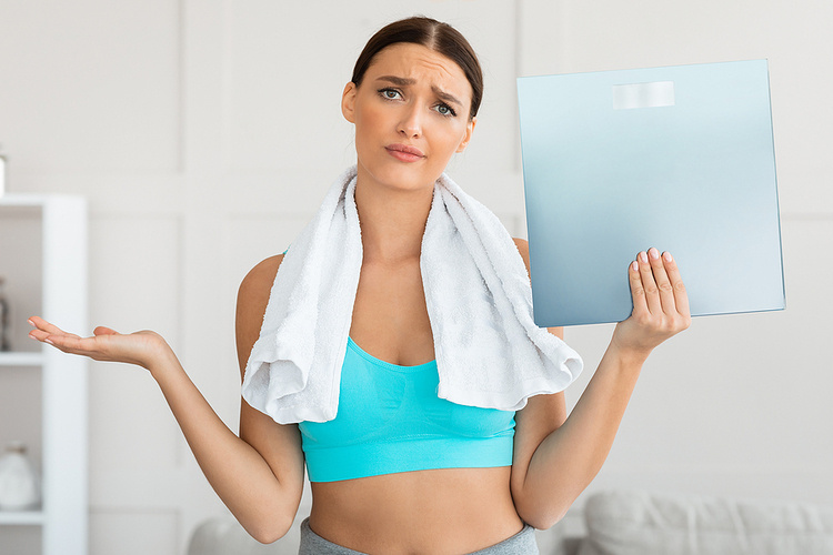 A woman holding a scale with a questioning look like she doesn't know what to do. She is wearing a towel around her neck and a blue shirt. Losing Weight Too Fast