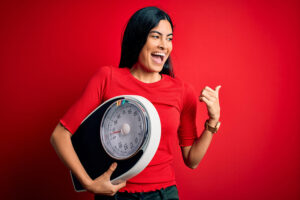 Young beautiful black-haired woman holding an analog bathroom scale. She's wearing a red shirt and jeans. There's a red background and she's giving a thumbs up and smiling. 7 Tips For Losing Weight Healthily