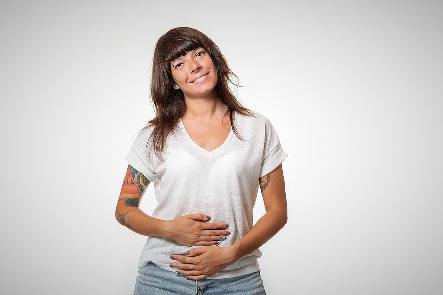 Fighting Against Your Body. A young brunette smiling. She's wearing a white plain t-shirt and blue jeans. She has tattoos on her arms and is holding her arms around her stomach.