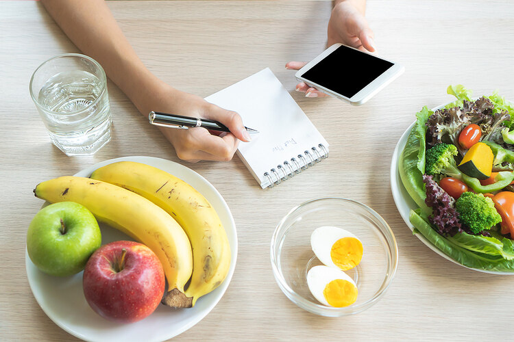 Woman using a smartphone to Eat Healthier by writing down a grocery list. she is holding a pen over a notepad. The notepad is on a table with plates of healthy foods, inclusing hardboiled eggs, fruit and a salad.