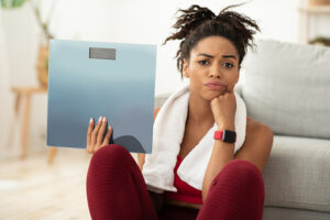 Upset Young Woman Holding a Bathroom Scale and Sitting On Floor against a sofa, Looking At Camera. She is wearing red workout clothes and has a white towel around her neck. Dieting, Over exercising, Worst Ways to Lose Weight, Weight Loss Failure.