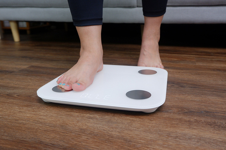 Photo of the feet of a Caucasian woman stepping onto a white, body fat measuring scale. The floor is dark brown wood and there is a gray couch in the background. The woman is wearing black leggings. Photo for Things I Wish I'd Known Before Losing Weight