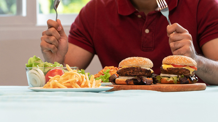 A picture of a man in a dark red t-shirt sitting at a table. He is holding a fork and spoon, and on the table is several plates of food. Included are french fries, two cheeseburgers, a small garden salad, and a plate of spaghetti with tomato sauce. Photo for a review of Overcoming Binge Eating by Dr. Christopher Fairburn