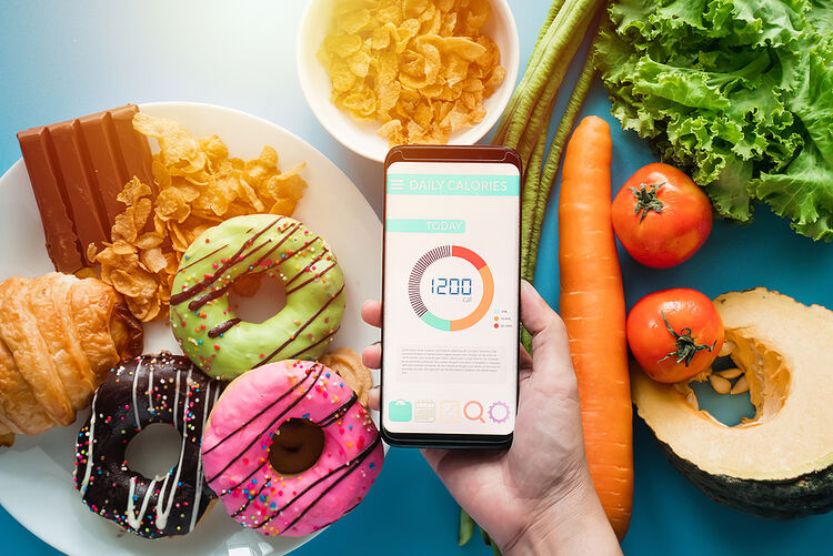 Image of a hand holding a smart phone displaying a calorie counting app. The app shows 1200 calories. In the background, there are several different types of food on a blue table. Shown are carrots, tomatoes, kale, string beans, a bowl of corn flakes and milk, a plate with donuts, a croissant, a Kitkat bar, and butter cookies. Photo for Diet Culture.
