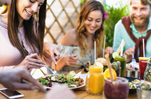 A photo of three young people sitting at a wooden table outside in the summer. The table is covered in healthy foods, including a plate of salad and jars filled with colorful fruit smoothies. The people are enjoying the food and smiling. Guide to Healthy Eating on a Budget