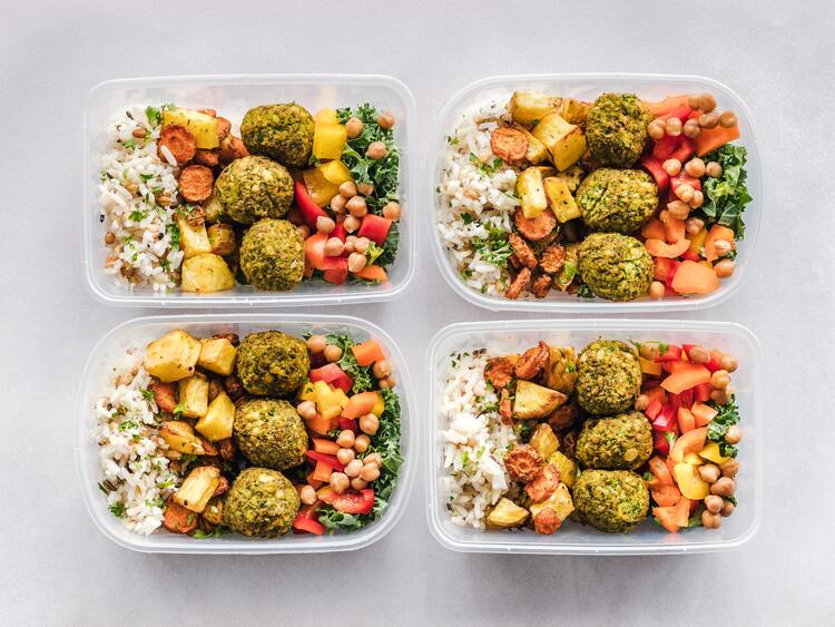 Photo of four plastic containers of food that has been meal prepped for lunches. They contain falafel, brown rice, salad with peppers, kale, and chickpeas, and roaster root vegetables. the containers are sitting side by side on a white table. Batching meals and healthy eating on a budget.