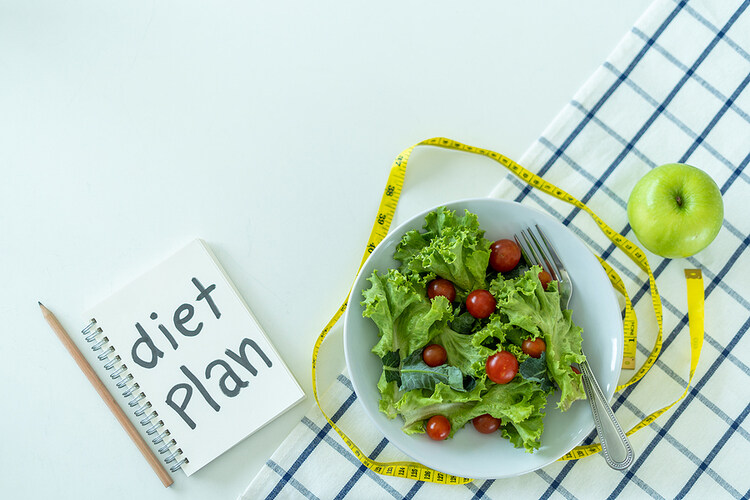 A photo displaying a restrictive dieting plan. There is a notebook that has the words diet plan written down. Nest to it, there is a garden salad with no dressing, surrounded with a yellow measuring tape and a green apple. They are sitting on a white table with a blue-striped, white tablecloth. Unsolicited diet advice Before Losing Weight 
