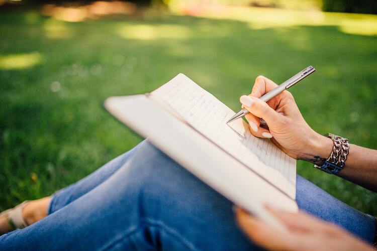 Photo of a woman sitting in a field under the shade of a tree. She is wearing blue jeans and a chain bracelet around her wrist. She holds a silver pen in her right hand over a notebook and is writing down her goals. mental health benefits of journaling