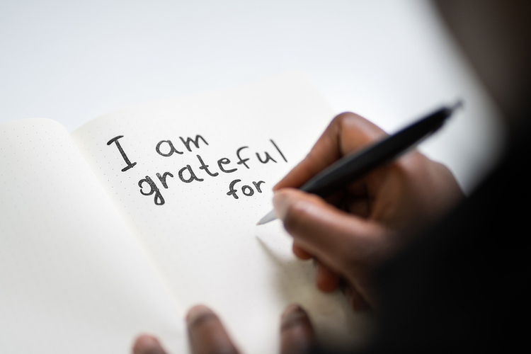 The photo shows someone's right hand holding a black pen over a notebook. It looks down at it from over their shoulder. The notebook is sitting on a white table. It has the words "I am grateful for" written on it in black ink. Photo for The Proven Mental Health Benefits of Journaling
