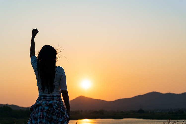 Photo for positive mental attitude. A picture of a woman with long, brown hair raising her fist to the sky. She has a white t-shirt on and a plaid sweater around her waist. The sky shows an orange sunset. In the distance, there's a lake with trees and mountains.