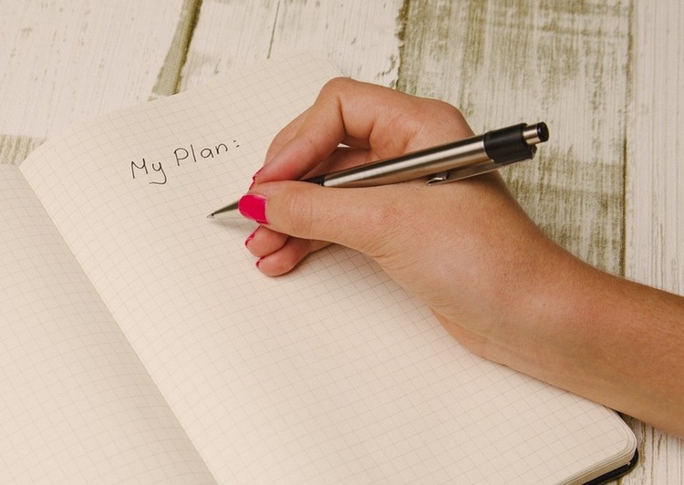 Photo of a hand writing a plan in a notebook. At the top of the paper the title my plan is written. The paper has grids drawn on it. The pen held in the hand is silver and black, and the person's fingernails are painted hot pink. The notebook is lying on a white, wooden table. Photo for Thinsanity by Glenn Mackintosh