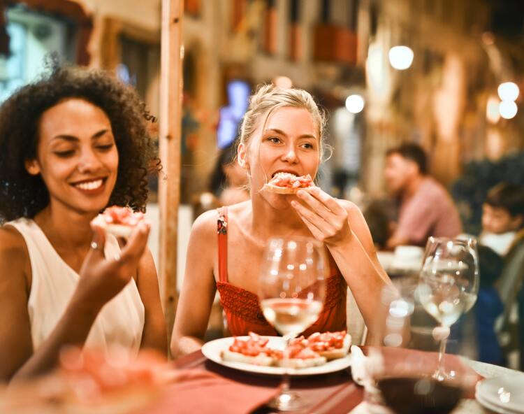 Photo of two young women sitting at a table with a plate of bruschetta, taking a bite out of it. There are wine glasses at the table filled with white wine. One woman has short, curly brown hair and the other has long, blond hair, tied back. There are other people sitting at tables in the background. Photo for hunger and fullness scale.