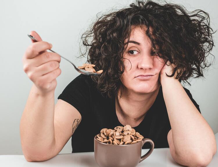Photo for 50 Things to Do Instead of Binge Eating. Photo of a woman with short, curly hair, wearing a black t-shirt, sitting at a white table. On the table, there's large brown mug filled with cereal. The woman is holding up a spoon filled with cereal and giving it a disgruntled look. The background is gray.