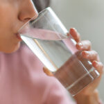 A photo of a young woman holding a glass of water to her mouth, taking a drink. She is wearing a light pink t-shirt. Picture for Stay Hydrated without Added Sugar Post