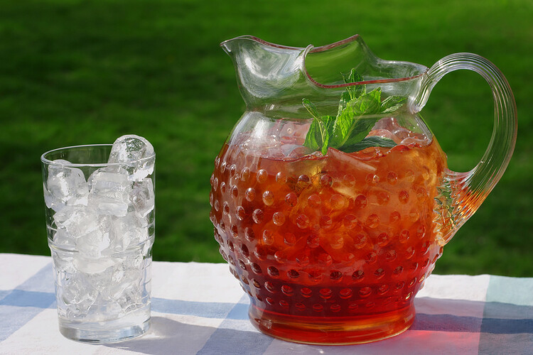 The picture shows a glass pitcher filled with iced tea and mint leaves next to a glass filled with ice. They are sitting on a table with a blue and white checkered tablecloth. The table is sitting in a grassy field. Photo for stay hydrated without added sugar post.