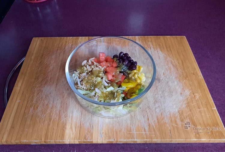 Photo of Vegetarian Burrito Bowls taken from above. The burrito bowl is in a glass bowl and has black beans, tomatoes, shredded lettuce, wild rice, corn, sliced rainbow peppers, and salsa verde. The bowl is sitting on a wooden cutting board.