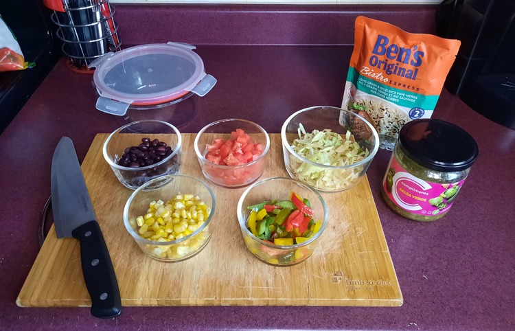 Photo of Vegan Burrito Bowls ingredients. There are several ingredients sitting in glass bowls on top of a wooden cutting board.  They include black beans, tomatoes, shredded lettuce, wild rice, corn, sliced rainbow peppers, and salsa verde. The cutting board has a knife on it and a glass container next to it, and is sitting on a purple counter.