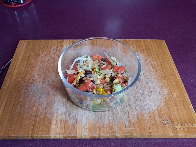 Photo of Vegetarian Burrito mixed up taken from above. The burrito bowl is in a glass bowl and has black beans, tomatoes, shredded lettuce, wild rice, corn, sliced rainbow peppers, and salsa verde. The bowl is sitting on a wooden cutting board on a purple counter.