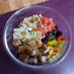 Photo of Burrito Bowls taken from above. The burrito bowl is in a glass bowl and has black beans, tomatoes, shredded lettuce, wild rice, corn, sliced rainbow peppers, and salsa verde. The bowl is sitting on a purple counter next to a wooden cutting board.